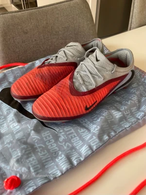 Nike fotbollsskor phantom 6 elite ag  - Snygga fotbollsskor från Nike i rött och ljusgrått med svarta detaljer. Skorna har en låg profil, snörning och ovandel i syntetmaterial för bra bollkontroll. Skorna är för konstgräs