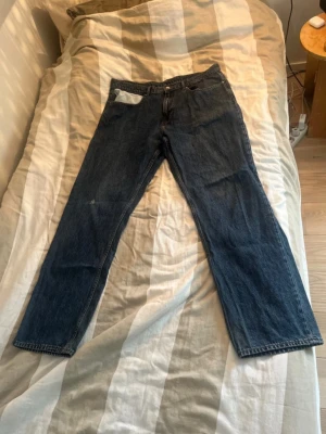 Mörkblå raka jeans - Säljer ett par mörkblå jeans med rak passform och klassisk femficksdesign. Jeansen har en enkel look utan synliga loggor och är tillverkade i denim. Liten skada på höger knä, annars helt felfria. Perfekta för dig som gillar en clean och tidlös stil.