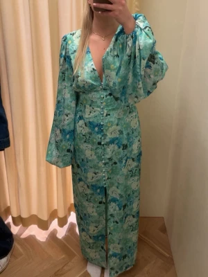 Blommig långklänning med v-ringning - Långklänning i gröna och blå toner med blommigt mönster. Klänningen har v-ringning, knappar framtill och vida långärmar. Perfekt för sommarens alla tillfällen.