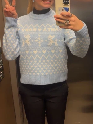 Ljusblå stickad tröja med mönster - Söt ljusblå stickad tröja med vita hjärtan, renar och texten 'SANTA BABY' över bröstet. Tröjan har rund hals, ribbade muddar och avslut. Perfekt för vintermys och julkänsla.