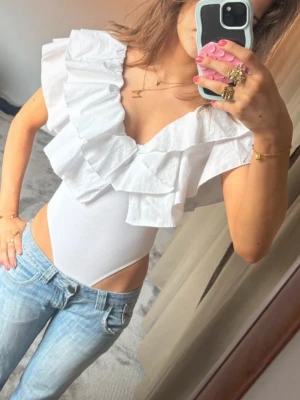 Vit Zara body - Säljer en vit body med stora volanger och djup urringning. Bodyn har breda axelpartier och är tillverkad i ett stretchigt material med volanger i bomullstyg. Perfekt att styla med jeans eller kjol för en trendig look.