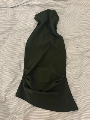 Mörkgrön halterneckklänning med bälte - Snygg mörkgrön halterneckklänning med markerad midja och tillhörande bälte. Klänningen har en figurnära passform upptill och lätt utsvängd kjol. Perfekt för dig som vill ha en stilren och trendig look.