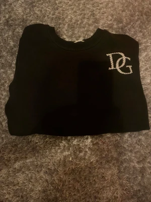 Svart Dolce & Gabbana hoodie - Säljer en svart hoodie från Dolce & Gabbana med DG-logga i vitt på bröstet. Klassisk rund halsringning och mjukt material som känns skönt mot huden. Perfekt för dig som vill ha en stilren och exklusiv look.