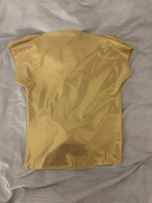 Guldig glittrig topp med kort ärm - Snygg guldig topp med glittrig finish och korta ärmar. Toppen har en enkel, rak passform och är tillverkad i ett glansigt syntetmaterial som ger en riktigt festlig look. Perfekt för dig som vill sticka ut med metallic-vibbar.