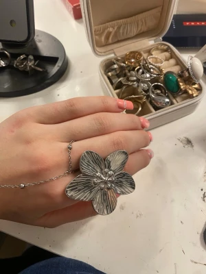 Stor silverfärgad blommring - Unik ring i silverfärg med stor blomformad design och detaljerade linjer på kronbladen. Ringen har en kedja som kopplar ihop fingret med en tunn ring, vilket ger en cool och trendig look. Perfekt för dig som vill sticka ut med dina accessoarer.