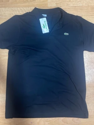 Svart pikétröja från Lacoste - Klassisk svart pikétröja från Lacoste med den ikoniska gröna krokodilloggan broderad på bröstet. Tröjan har korta ärmar, krage och knappar vid halsen. Tillverkad i bomull för en bekväm och stilren look.
