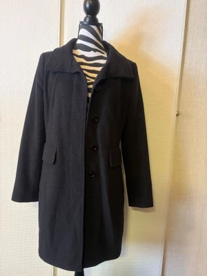 Svart klassisk ullrock - Stilren svart ullrock med knäppning framtill och två fickor med lock. Rocken har en klassisk krage och rak passform som ger en tidlös look. Perfekt för kyligare dagar och enkel att matcha med olika outfits.