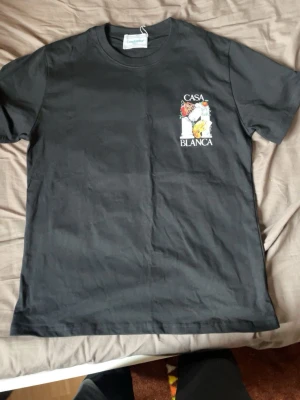 Svart t-shirt från Casablanca - Svart t-shirt från Casablanca med färgglatt tryck av pelare, schackbräde och frukt samt texten 'Casa Blanca' både fram och bak. Klassisk rund hals och korta ärmar. Perfekt för dig som gillar streetwear med unik design.