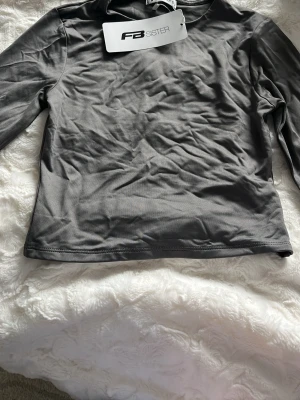 Grå långärmad topp från FB Sister - En enkel och stilren grå långärmad topp från FB Sister. Toppen har en rund halsringning och är tillverkad i ett mjukt syntetmaterial som känns bekvämt mot huden. Perfekt att matcha med jeans eller kjol för en clean look.