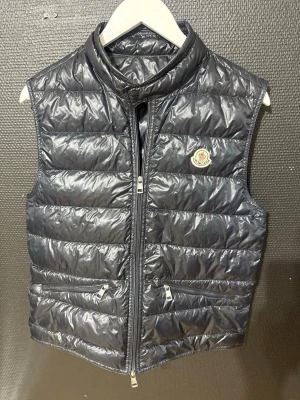 Moncler gui väst - Snygg svart dunväst från Moncler med quiltad design och hög krage. Västen har två fickor med dragkedja och Moncler-logga på bröstet. Materialet är glansigt och lätt, perfekt för lager-på-lager och coola outfits. Storlek (1)