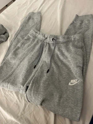 Grå mjukisbyxor från Nike - Snygga grå mjukisbyxor från Nike med dragsko i midjan och klassisk logga på benet. Byxorna har resår i midjan och vid bensluten, samt mjukt och skönt material som passar perfekt för chill eller träning.