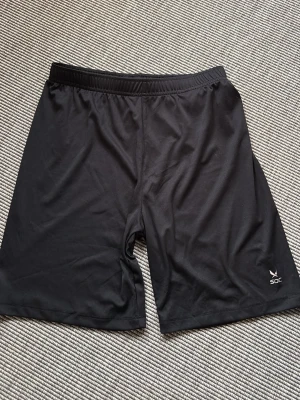 SOC - Svarta junior träningsshorts från SOC - Svarta junior träningsshorts från SOC med elastisk midja och snörning. Tillverkade i ett lätt och smidigt polyestermaterial som andas. Perfekta för sport eller gym. Klassisk design utan mönster och med diskret logga på benet.