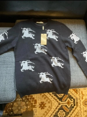 Svart stickad tröja  - Snygg svart stickad tröja från Burberry med vita riddarmotiv över hela plagget. Tröjan har rund halsringning och ribbade muddar vid ärmslut och nederkant. Perfekt för dig som gillar klassiska designer med en twist. Priset diskuteras