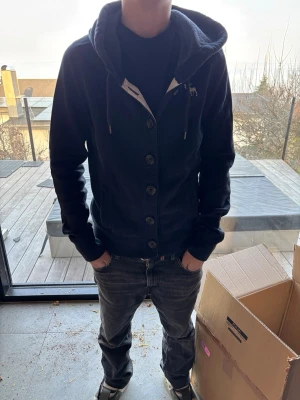 Mörkblå hoodie med knappar - Snygg mörkblå hoodie med knappar framtill och dragsko i huvan. Hoodien har en liten broderad logga på bröstet och ribbade muddar vid ärmslut och nederkant. Perfekt för en avslappnad och stilren look. Storlek m men passar i stl xs