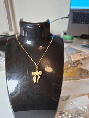 Guldigt halsband med rosett och pärla - Snyggt halsband med tunn guldfärgad kedja och ett hängsmycke i form av en rosett dekorerad med små gnistrande stenar och en vit pärla i mitten. Perfekt accessoar för dig som gillar detaljer och vill sticka ut lite extra.