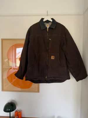 Brun Carhartt jacka med dragkedja - Mörkbrun Carhartt jacka med klassisk krage i kontrastfärg och grov dragkedja framtill. Två stora framfickor och Carhartt-logga på bröstet. Jackan är fodrad med mjuk fleece och har en loose passform, perfekt för kalla dagar.