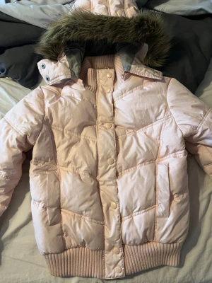Y2k jacka  - Ljusrosa pufferjacka med avtagbar huva och fuskpälsdetalj. Jackan har ribbade muddar vid ärmslut och nederkant, tryckknappar framtill och två sidofickor. Snygg quiltad design och varm vaddering, perfekt för kalla dagar. Priset kan diskuteras, passar som en storlek S!!
