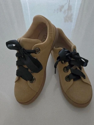 Beige Sneakers i Mocka med Svarta Band - Snygga TEXTO dam-sneakers “Made in Italy” i beige mocka-liknande material. Skorna har breda svarta band som skosnören och metallöljetter, vilket ger ett elegant och modernt utseende. Den neutrala färgen gör dem lätta att matcha med jeans, kjol eller andra vardagskläder.