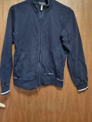 Mörkblå zip hoodie från Champion - Mörkblå hoodie från Champion med dragkedja framtill och två fickor. Tröjan har vita detaljer vid muddar och nederkant samt en liten Champion-logga på vänster sida. Tillverkad i mjukt bomullsmaterial och har en klassisk, enkel design utan huva.