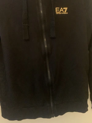 Svart hoodie från EA7 Emporio Armani - Snygg svart hoodie från EA7 Emporio Armani med dragkedja framtill och guldfärgad logga på bröstet. Hoodien har justerbar huva med snören och ribbade muddar. Tillverkad i mjukt bomullsmaterial som känns skönt mot huden.