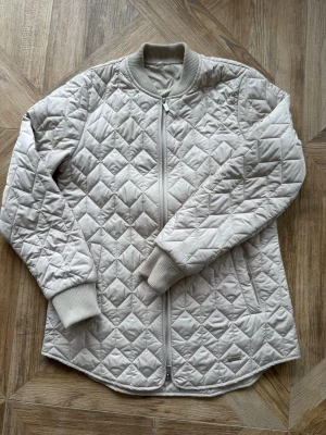 Beige quiltad vårjacka Ilse Jacobsen - Snygg beige quiltad jacka med dragkedja framtill och ribbade muddar vid ärmslut och krage. Jackan har en avslappnad passform och två sidofickor. Perfekt för lager på lager och ger en clean look till din outfit.