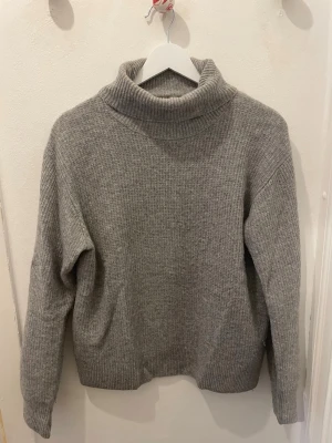 Grå stickad polotröja - Säljer en enkel och stilren grå stickad polotröja med ribbade muddar och hög krage. Tröjan har lång ärm och är gjord i mjukt material som passar perfekt när det är kyligt ute. Perfekt för dig som gillar klassiska och bekväma plagg.