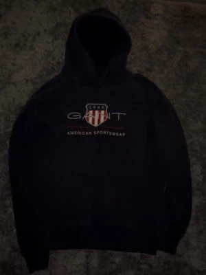 Gant hoodie - Svart hoodie från Gant med huva och stort tryck på bröstet där det står 'GANT 1949 American Sportswear' samt ett rött och vitt sköldmärke. Klassisk passform och ribbade muddar. Perfekt för en avslappnad och sportig stil.