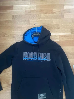 Svart HOODRICH hoodie med blå detaljer - Svart hoodie från HOODRICH med blå och grå logga framtill och blå insida på huvan. I storlek S.Stor magficka och tryck även på ryggen. Snygg streetwear-stil med dragsko i huvan och ribbade muddar. Perfekt för dig som gillar urban mode.