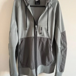 Nike Air Max grå hoodie med dragkedja - Snygg hoodie från Nike Air Max i två nyanser av grått. Modellen har hel dragkedja framtill, huva med svarta dragsnören och fickor på sidorna. Tillverkad i mjukt bomullsmaterial och har detaljerade sömmar samt Air Max-logga på bröstet.Priset kan diskuteras vid snabbaffär.