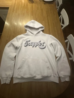 Vit hoodie från Trapper med brodyr - Säljer en vit hoodie från MadeByTrappers med stor broderad logga i grått och vitt på bröstet. Hoodien har klassisk känguruficka, ribbade muddar och luva. Perfekt för chill dagar och streetwear-stil. Materialet känns mjukt och bekvämt. Företaget som spålde denna hoodie har tyvärr gott i konkan. Finns två små fläckar som ni ser på bild 3 och 4 