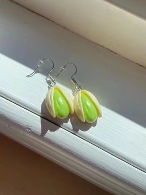 Örhängen med pistage-nöt design - Unika örhängen formade som pistagenötter i beige och ljusgrönt. De har en realistisk look och är tillverkade i plast eller polymerlera. Perfekta för dig som gillar roliga och annorlunda accessoarer.