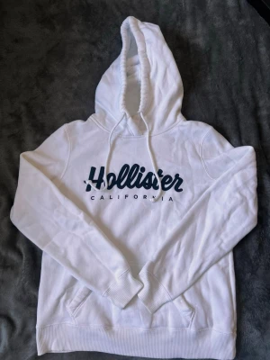 Vit hoodie från Hollister - Säljer en vit hoodie från Hollister med stor logga i mörkblått på bröstet och texten 'California' under. Hoodien har en klassisk känguruficka, ribbade muddar och dragsko i huvan. Mjuk insida i fleece och avslappnad passform. Perfekt för chill dagar.
