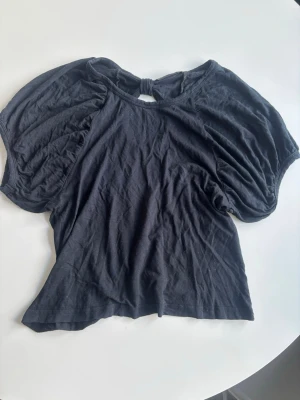 Svart topp med puffärmar och knut - Trendig svart topp med puffiga kortärmar och en snygg knutdetalj i nacken som skapar en liten öppning i ryggen. Toppen har en loose passform och är tillverkad i mjuk bomull.