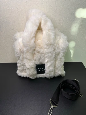 Vit fluffig handväska med axelrem - Supersöt vit handväska i mjuk fuskpäls med två handtag och avtagbar svart axelrem. Väskan har en fyrkantig form och en svart patch framtill med texten 'THE TOTE BAG'. Perfekt för dig som vill sticka ut med en mysig accessoar.