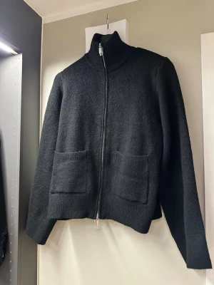 Svart zip-kofta med fickor - Mysig svart kofta med dragkedja framtill och två stora fickor. Rak passform och hög krage ger en clean look. Perfekt att slänga på sig över en t-shirt eller hoodie. Materialet ser ut att vara ull eller ullmix för extra värme.   Knitted pocket cardigan, slutsåld på ginas hemsida!🌸