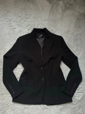 Svart kavaj från HM - Svart shaped kavaj från HM med två knappar framtill och klassisk krage. Storlek 38 men sitter som en 36