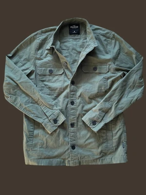 Olivgrön overshirt från Hollister - Stilren olivgrön overshirt från Hollister med klassisk krage, två bröstfickor med knappar och svarta knappar framtill. Tillverkad i bomull för en skön känsla och perfekt lager på lager. Enkel och clean design som funkar till det mesta. Modellen är 185 och väger 65.