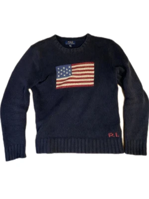Tröja Polo Ralph Lauren USA - Mörkblå stickad tröja från Polo Ralph Lauren med amerikansk flagga framtill och RL-broderi vid nederkanten. Tröjan har rund halsringning och ribbstickade muddar. Perfekt för dig som gillar klassisk och preppy stil. Väldigt bra skick!