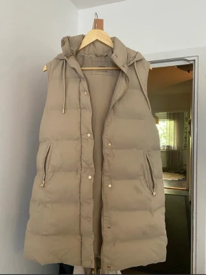 Beige lång väst med huva från Zara - Snygg beige lång väst från Zara med dragkedja och tryckknappar framtill. Västen har två fickor med dragkedja och en mysig huva med dragsko. Perfekt för lager-på-lager och chill höststil.