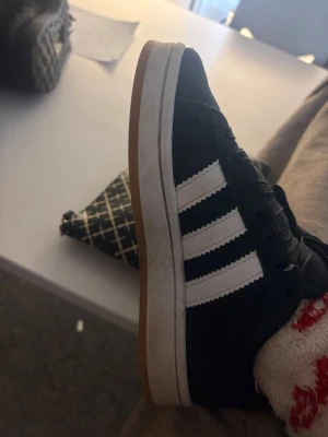 Svarta Adidas sneakers med vita ränder - Klassiska svarta Adidas sneakers med tre vita ränder på sidan och brun gummisula. Skon har en låg profil och ovandelen är i mocka för en stilren och tidlös look. Perfekt för dig som gillar streetwear och sportig stil.