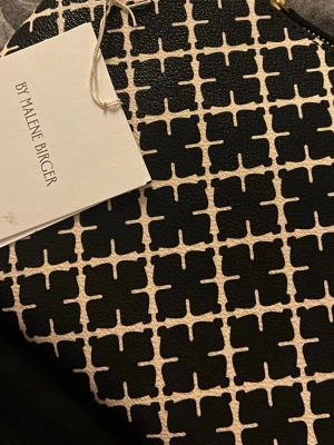 Kuvertväska By Malene Birger svart/vit - Snygg kuvertväska från By Malene Birger i svart och vitt med det klassiska grafiska mönstret. Väskan har dragkedja upptill och är gjord i ett tåligt konstlädermaterial. Perfekt accessoar för att lyfta din outfit.