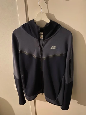 Mörkblå Nike Tech hoodie med dragkedja - Snygg mörkblå hoodie från Nike med hel dragkedja framtill och huva. Har en diskret logga på bröstet och en ficka med dragkedja på ärmen. Tillverkad i ett mjukt och stretchigt material som passar perfekt till streetwear-stilen.