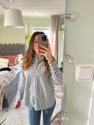 Randig skjorta  - Snygg skjorta med vita och gråa vertikala ränder. Skjortan har klassisk krage, knäppning framtill och långa ärmar. Passformen är loose och materialet känns som bomull, perfekt för en avslappnad stil.