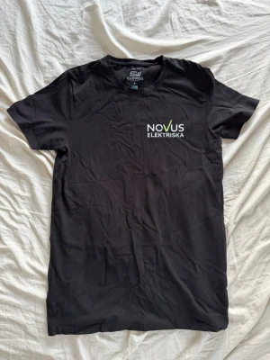 Svart t-shirt Novus Elektriska - Svart t-shirt från Southwest med trycket 'Novus Elektriska' på bröstet. Klassisk rund hals och korta ärmar. Materialet är mjuk bomull som känns skönt mot huden. Perfekt för dig som gillar enkel och stilren design med en touch av företagsprofil.