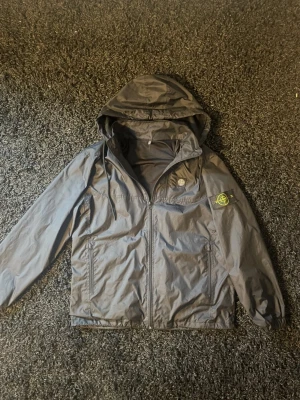 Svart vindjacka från Stone Island - Snygg svart vindjacka från Stone Island med huva och dragkedja framtill. Jackan har en bröstficka med dragkedja och klassisk Stone Island-logga på ärmen. Tillverkad i lätt och slitstarkt material, perfekt för blåsiga dagar.