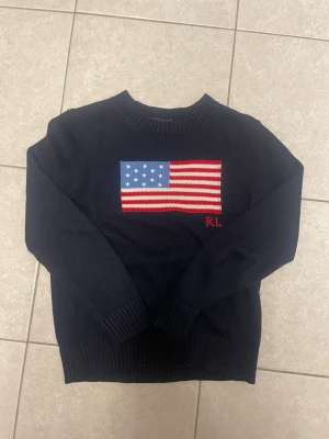 Mörkblå stickad tröja Ralph Lauren - Mörkblå stickad tröja från Ralph Lauren med amerikansk flagga i rött, vitt och blått på bröstet samt RL-broderi. Tröjan har rund halsringning och ribbstickade muddar vid ärmslut och nederkant. Perfekt för en stilren och klassisk look. Storlekslappen har åkt bort men passar xs