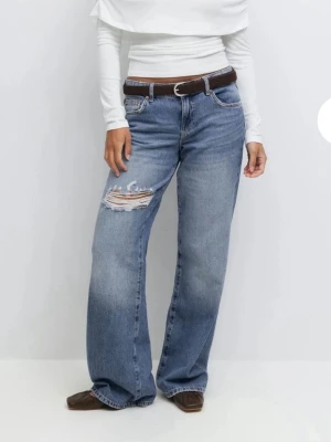 Low rise raka jeans med hål - Säljer ett par blåa raka jeans med låg midja och en stor sliten detalj på vänster lår. Jeansen har klassisk femficksmodell och är tillverkade i denim. Perfekta för en avslappnad och trendig look. Aldrig använt prislapp på‼️