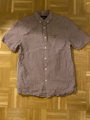 Rutig kortärmad skjorta från Lyle & Scott - Kortärmad skjorta från Lyle & Scott i brunt och vitt rutigt mönster. Klassisk button-down krage och gul logga på bröstet. Tillverkad i bomull och har knappar framtill. Perfekt för en avslappnad och stilren look.