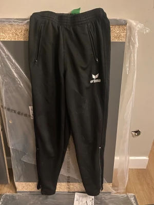 Svarta träningsbyxor från Erima - Säljer ett par svarta träningsbyxor från Erima med dragkedjor vid fickorna och bensluten. Byxorna har resår i midjan och en enkel, sportig design med logga på låret. Perfekta för träning eller chill.