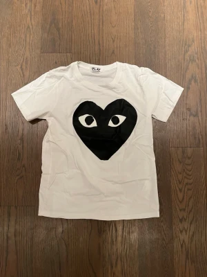 Vit Comme des Garçons Play t-shirt - Vit t-shirt från Comme des Garçons Play med det ikoniska svarta hjärtat med ögon tryckt på bröstet. Klassisk rund hals och korta ärmar. Perfekt för dig som gillar streetwear och vill ha något stilrent men ändå lekfullt.